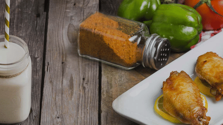 A jar of milk with a paper straw sits on a rustic wooden table next to a plate of fried food on lemon slices, with spice and peppers.
