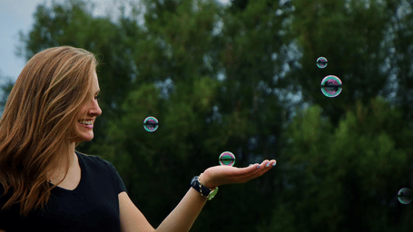 A smiling woman with long auburn hair holding her hand out towards floating bubbles outdoors.