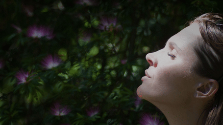A woman in profile, eyes closed, takes a deep breath surrounded by dark green and purple foliage.