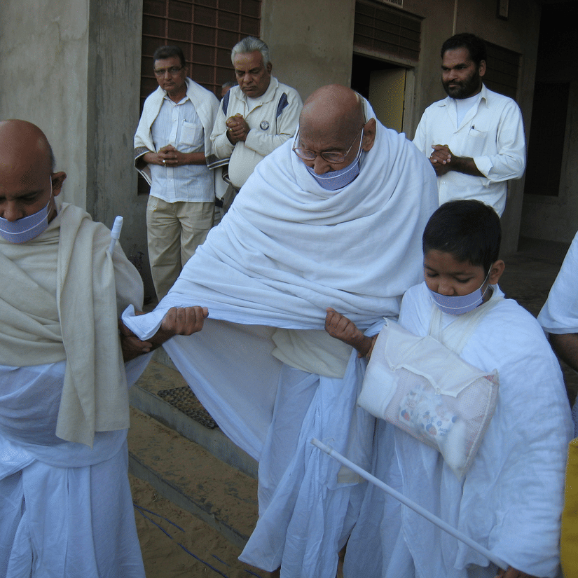 A group of Jain monks dressed in traditional white robes, with several individuals wearing white cloth masks (mukhavastrika) over their mouths and noses.