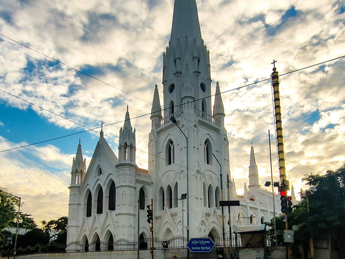 A low-angle shot of a white cathedral with a tall, pointed spire set against a dramatic sky with clouds.