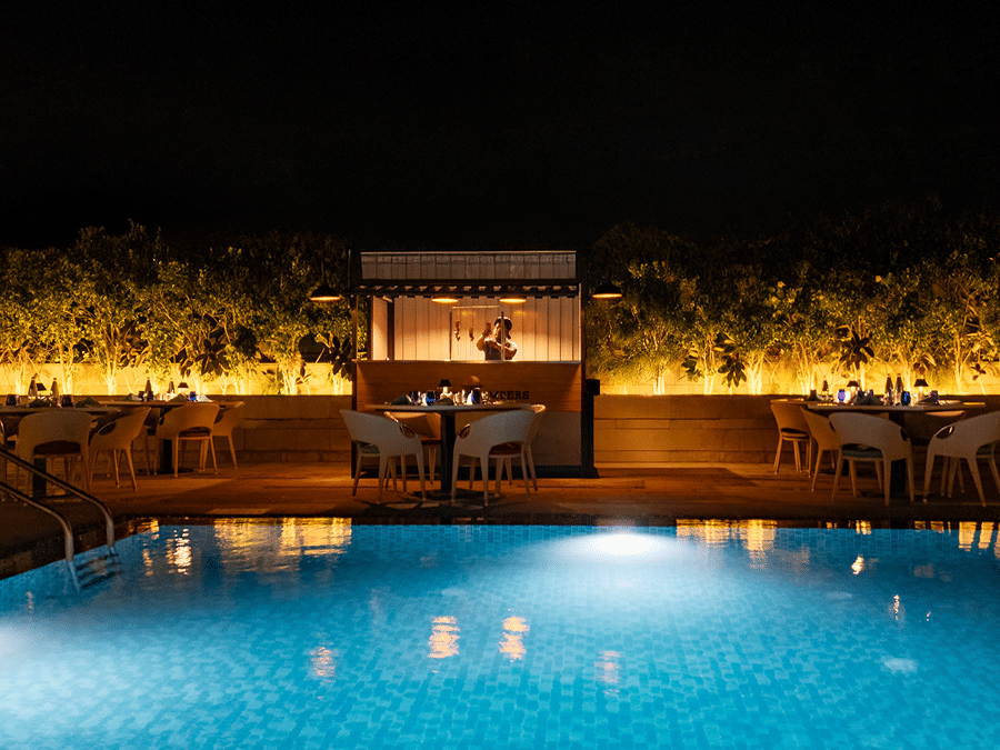A tranquil nighttime view of the hotel's outdoor swimming pool, lit up with glowing lights, with a seating area surrounded by plants at The Residency Towers Rameswaram.