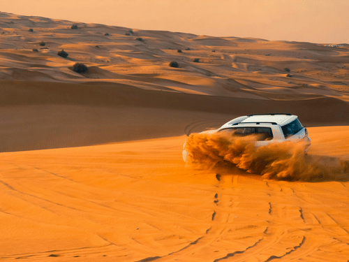 A white SUV kicks up a large cloud of sand while driving fast across rolling orange desert sand dunes at sunset.