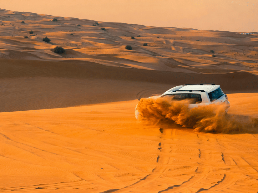 A white SUV kicks up a large cloud of sand while driving fast across rolling orange desert sand dunes at sunset.