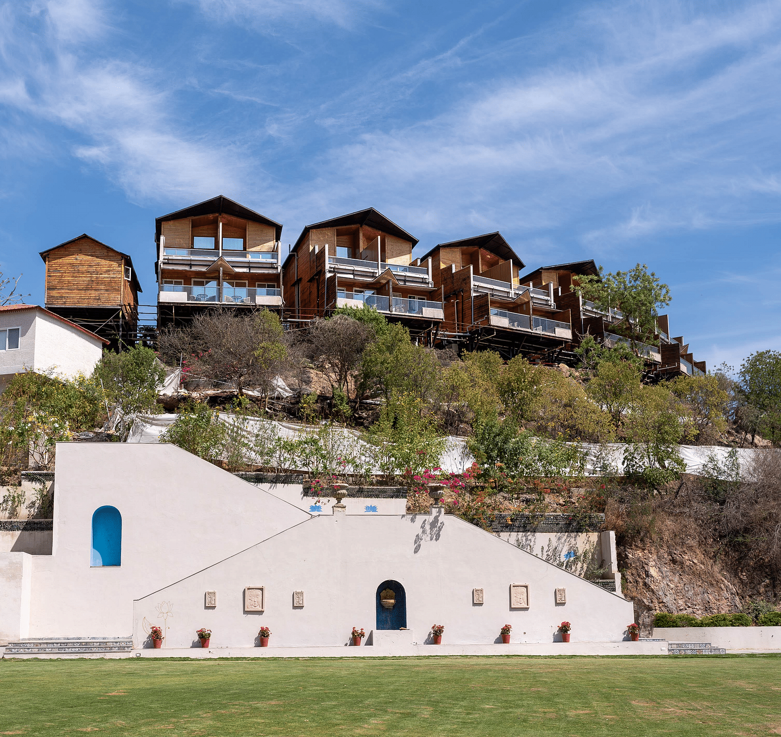 Buildings with accents at the base of a hill, leading to a cluster of wooden houses on the hilltop.- Bamboo Saa Resort & Spa, Udaipur