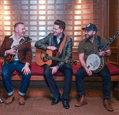 Four men sit on a red bench with musical instruments: a violin, mandolin, guitar, and banjo. They are smiling warmly, indicating camaraderie.