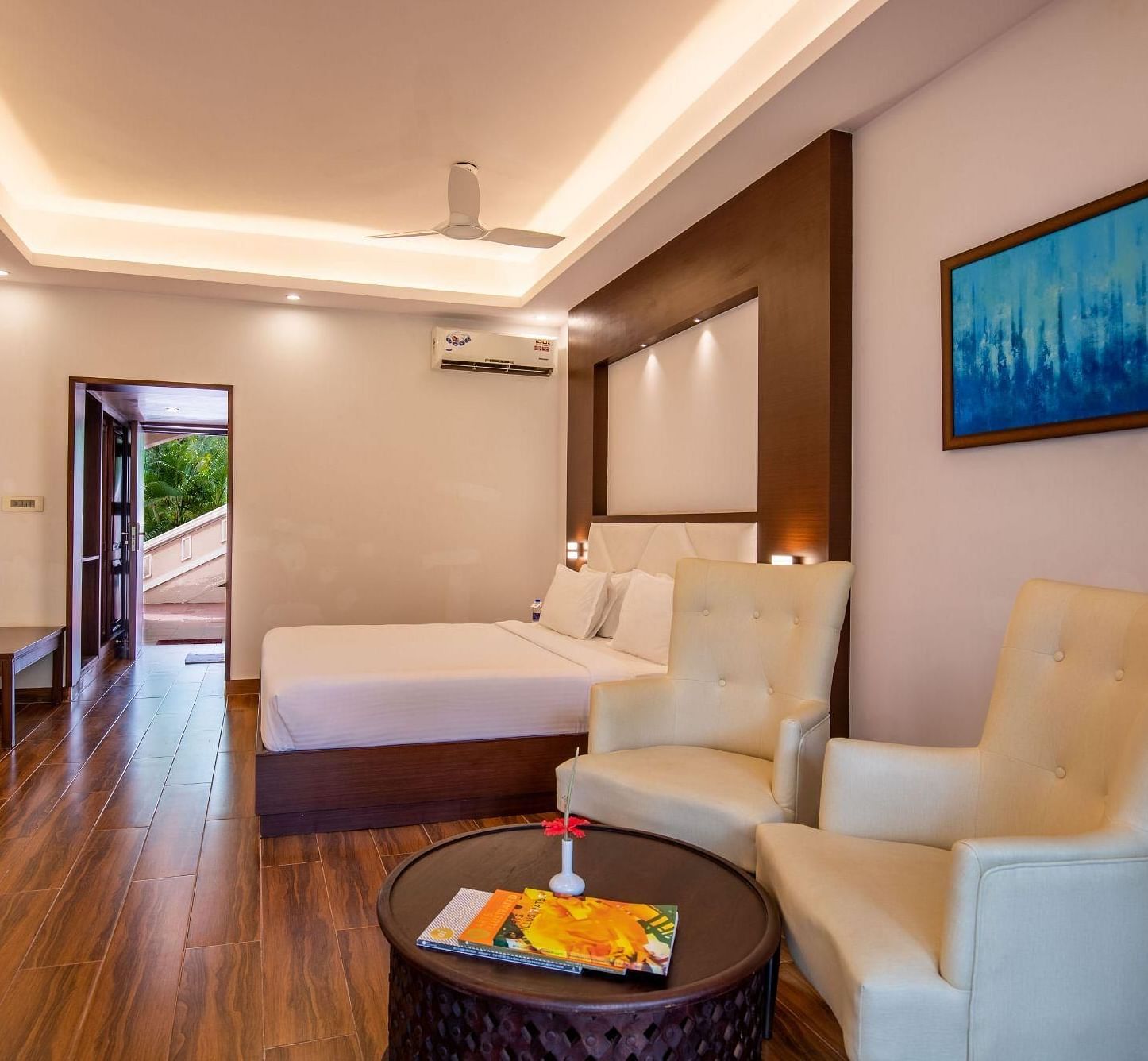 A contemporary hotel bedroom at Avana Resort with a large bed, two armchairs, and a round coffee table, featuring wooden floors and a balcony. 