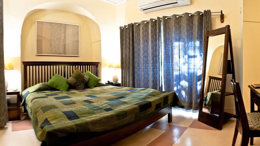 Bedroom with a bed featuring a patchwork quilt, green pillows, bedside tables with lamps, a full-length mirror, and curtains at Tijara Fort-Palace, Alwar.