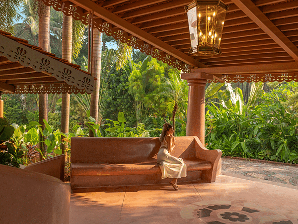 A girl relaxing on a sofa in the spacious outdoor seating area at Heritage Village Resorts & Spa, Goa, with a chandelier hanging overhead and large trees in the background