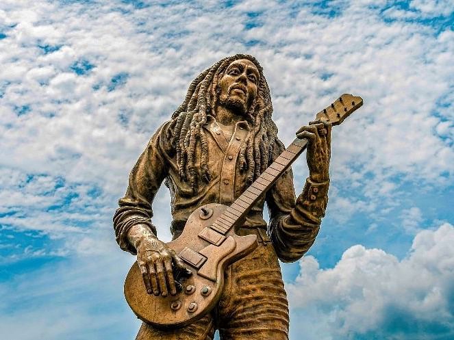 A brass statue of Bob Marley holding a guitar with a clouded blue sky in the background