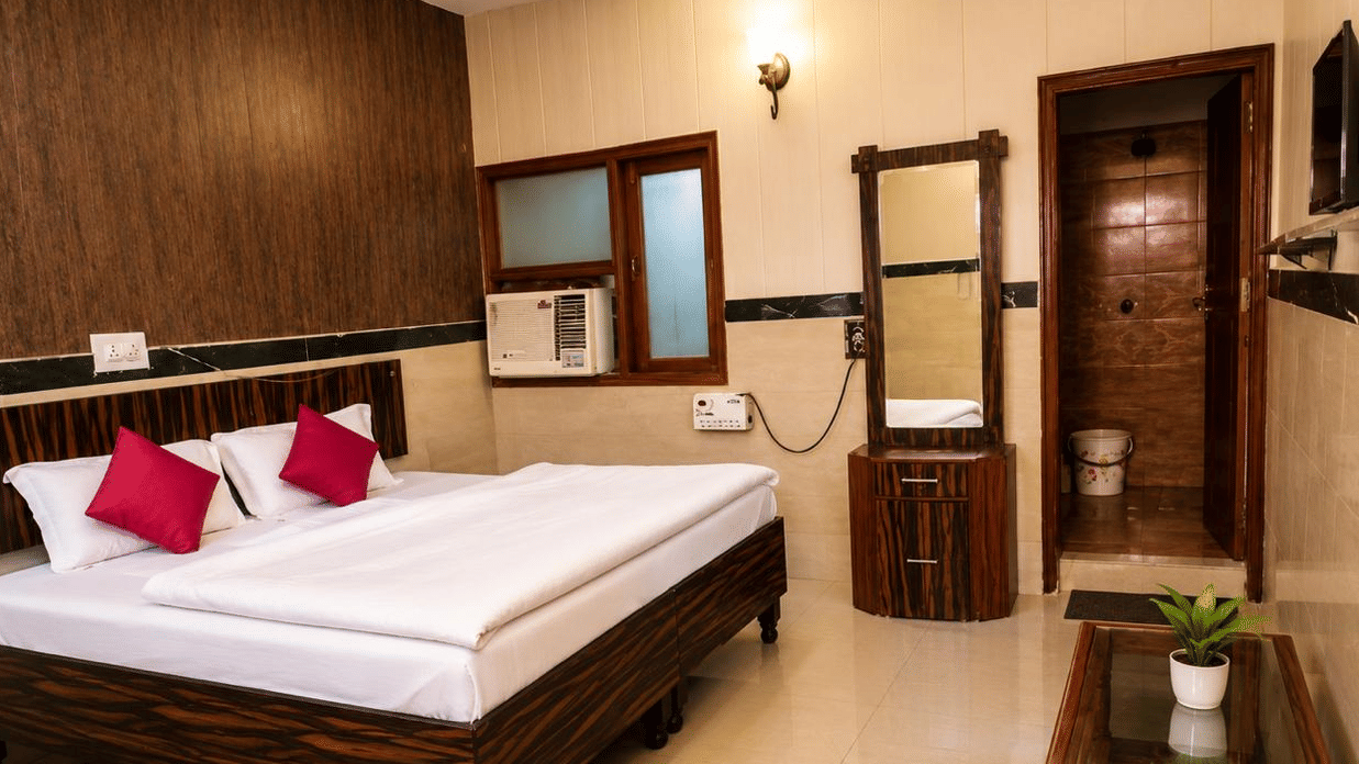 Bedroom interior of Hotel Goverdhan, Agra, featuring king size bed with white linen, 2 pillows, wooden headboard, window air conditioner, dressing table, mirror, table, and plant.