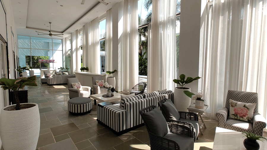 Bright hallway at The Mansion House resort with grey armchairs, striped ottomans, white conical planters, floor-to-ceiling sheer curtains, and polished light stone floor