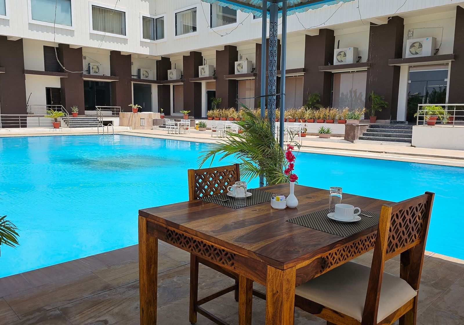 Shot of an outdoor dining area at the Bamboo Saa Mulberry Resort, Pushkar, featuring a wooden table and two chairs set beside a bright blue swimming pool.
