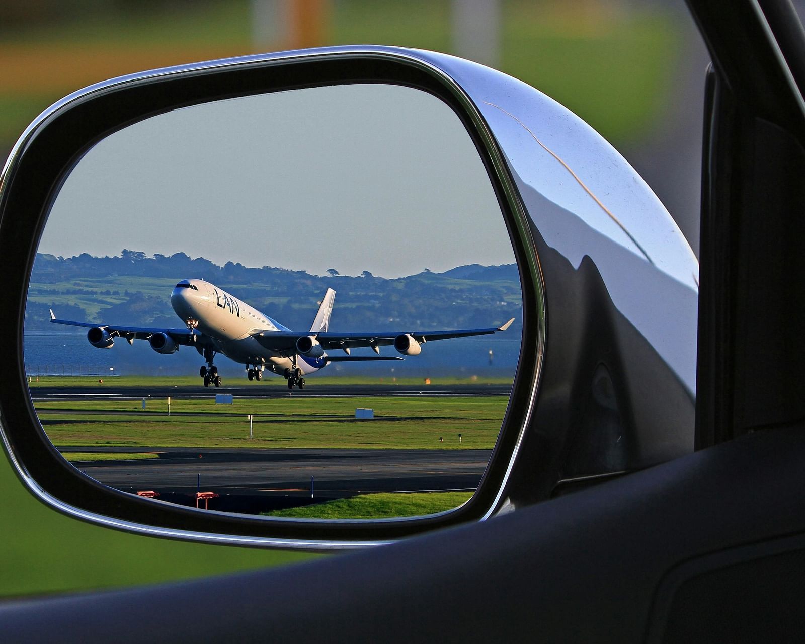 A car's side mirror reflecting an airplane on a runway at an airport.