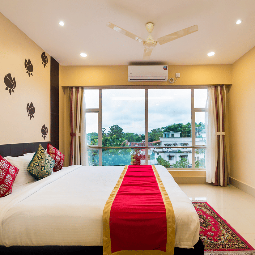 A neatly arranged bed with stylish interior, featuring a wall-mounted TV and a glass window offering a scenic view at Central Courtyard Boutique Hotel, Siliguri