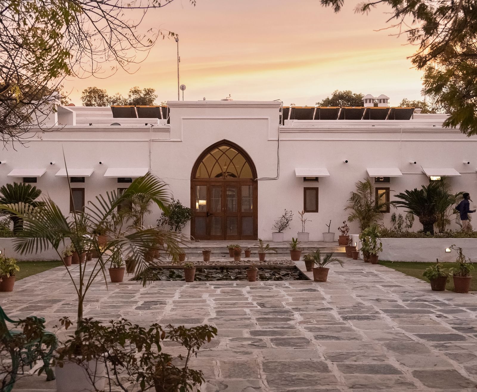 A white, single-story building with an arched doorway and paved courtyard under a warm sky at sunrise or sunset- Shahpura Bagh.