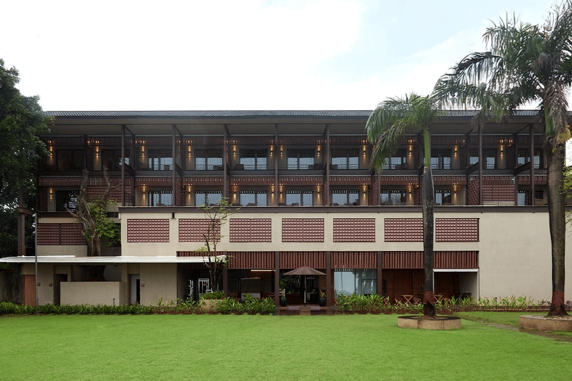 Exterior of a modern hotel building with wooden screens and balconies, fronted by a large, well-maintained green lawn and tall palm trees.