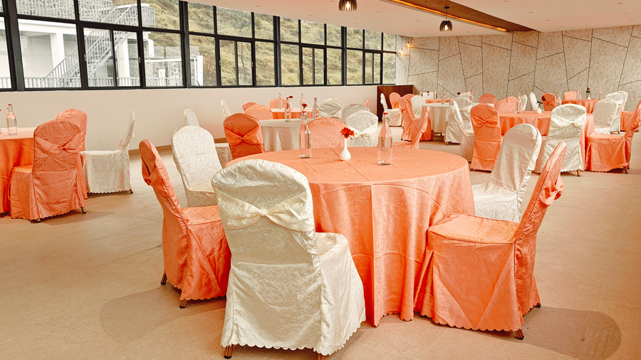 A close view of round tables with peach linens and white chair covers in a hall with windows at Bliss Casa Mila By Bergamont Hotels, Kodaikanal.
