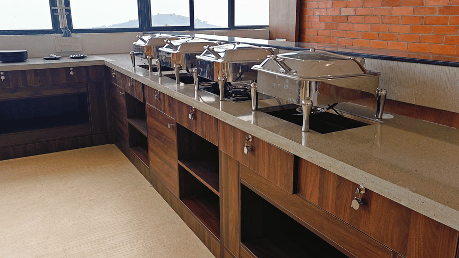 Silver chafing dishes are placed on a long wooden buffet counter with a brick wall at Bliss Casa Mila By Bergamont Hotels, Kodaikanal.