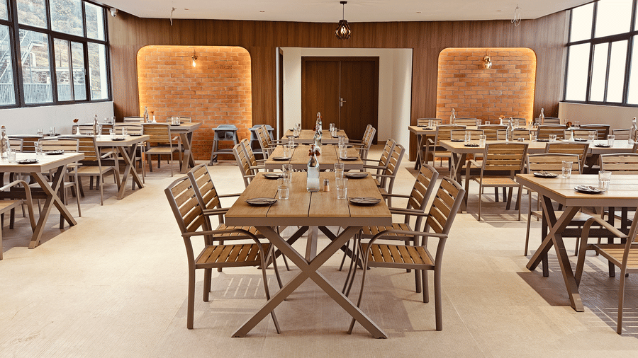 A symmetrical view of a dining hall showing wooden tables and chairs under hanging lights at Bliss Casa Mila By Bergamont Hotels, Kodaikanal.