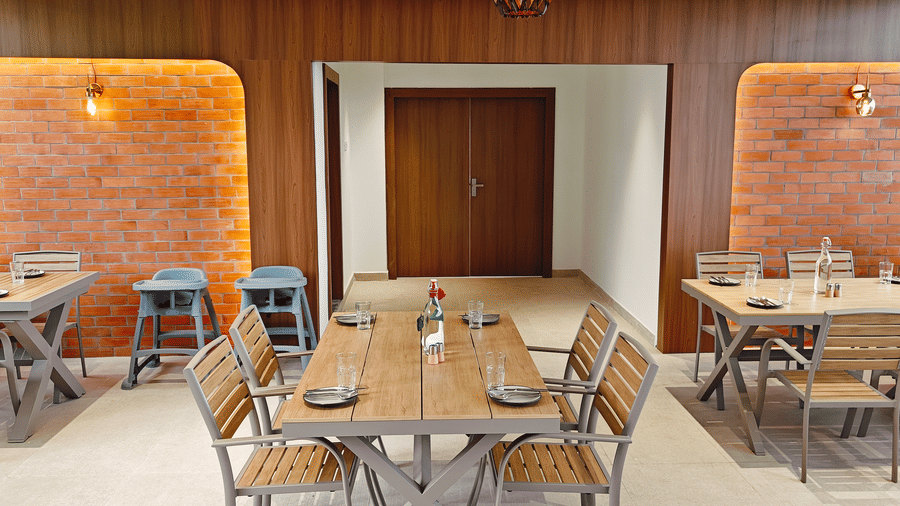 Wooden dining tables are arranged between 2 glowing brick wall alcoves and a doorway at Bliss Casa Mila By Bergamont Hotels, Kodaikanal.