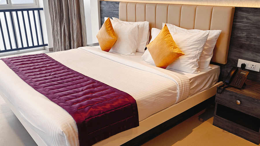A large bed with white linens, yellow cushions, and a purple runner sits next to a dark wood side table at Bliss Casa Mila By Bergamont Hotels, Kodaikanal.