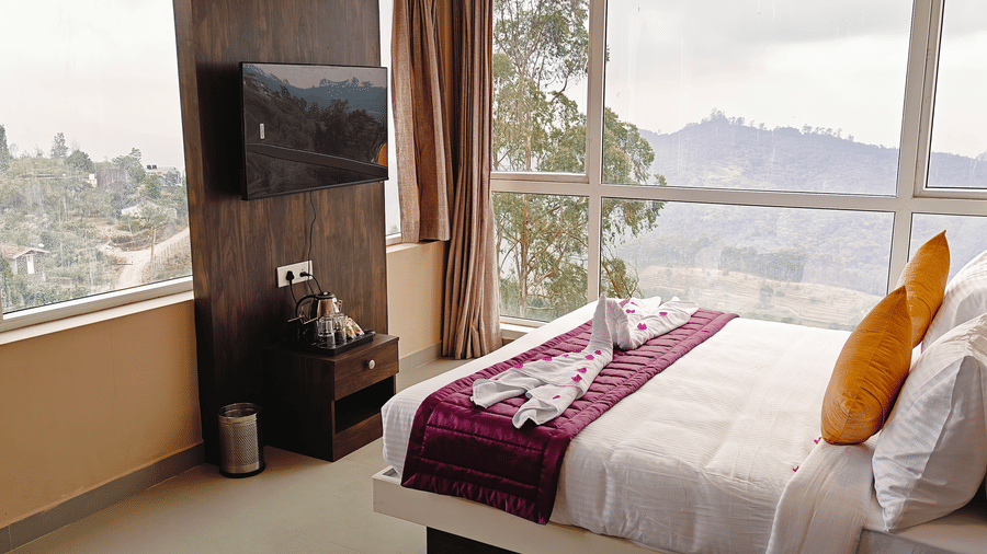 A bedroom with a television and a bed placed next to a large window showing green hills at Bliss Casa Mila By Bergamont Hotels, Kodaikanal.