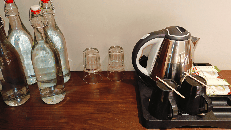A metal electric kettle, 4 glass bottles of water, and 2 black mugs on a wooden surface at Bliss Casa Mila By Bergamont Hotels, Kodaikanal.