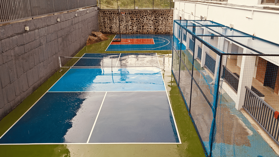 View of 2 blue sports courts and a fenced area next to a stone wall and white building at Bliss Casa Mila By Bergamont Hotels, Kodaikanal.