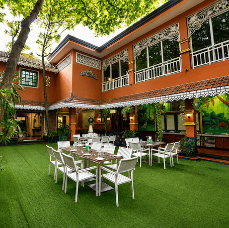 Aangan courtyard restaurant with white tables and chairs on a lawn, terracotta building with fretwork trim and a painted mural at MAYFAIR Garden, Rourkela.