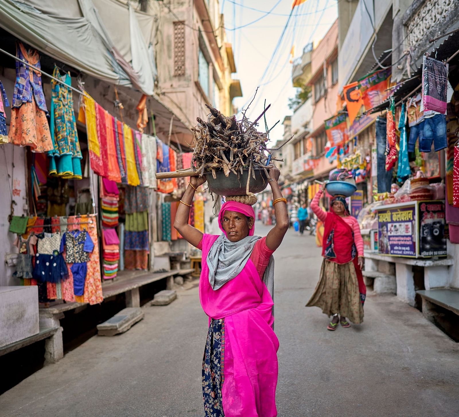 image of a woman wearing a pink half saree carrying firewood over her head through the streets in rajasthan