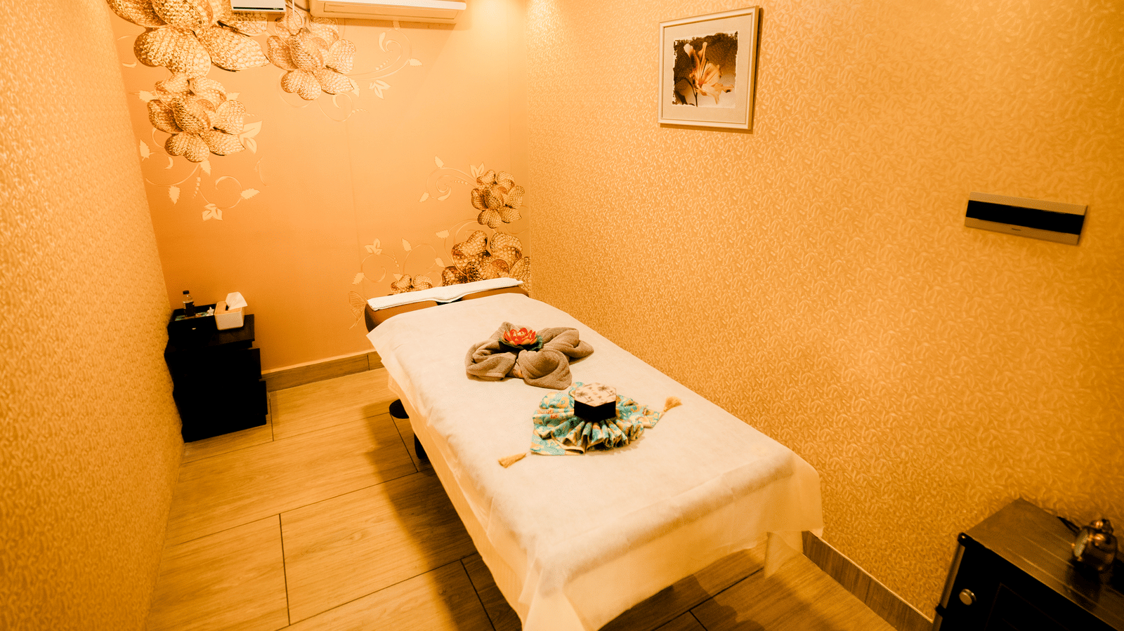 Spa treatment room with a massage bed, towels folded into a flower, gold floral wall decor and framed art at Hasta Spa, Ilara Hotels & Spa, Tada.