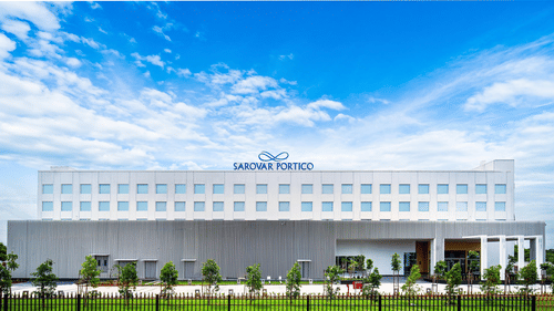 Exterior view of Sarovar Portico, Sriperumbudur, with signage on the top, set against a bright blue sky with scattered clouds and greenery in the foreground.