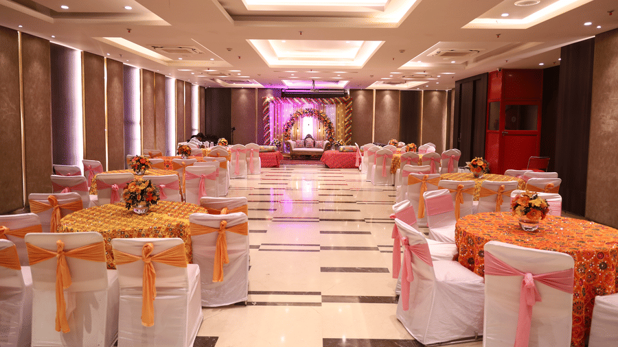 A banquet hall with round dining tables, chair covers with bows, floral centerpieces, and a decorated stage setup for a wedding ceremony at Hotel Saket 27.