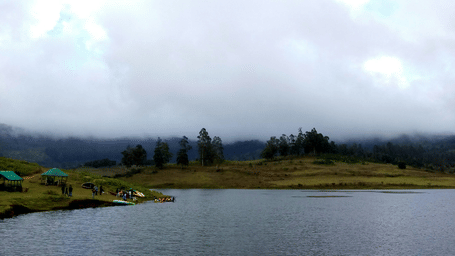 Calm lake surrounded by misty hills and cloudy skies, reflecting a serene natural landscape.