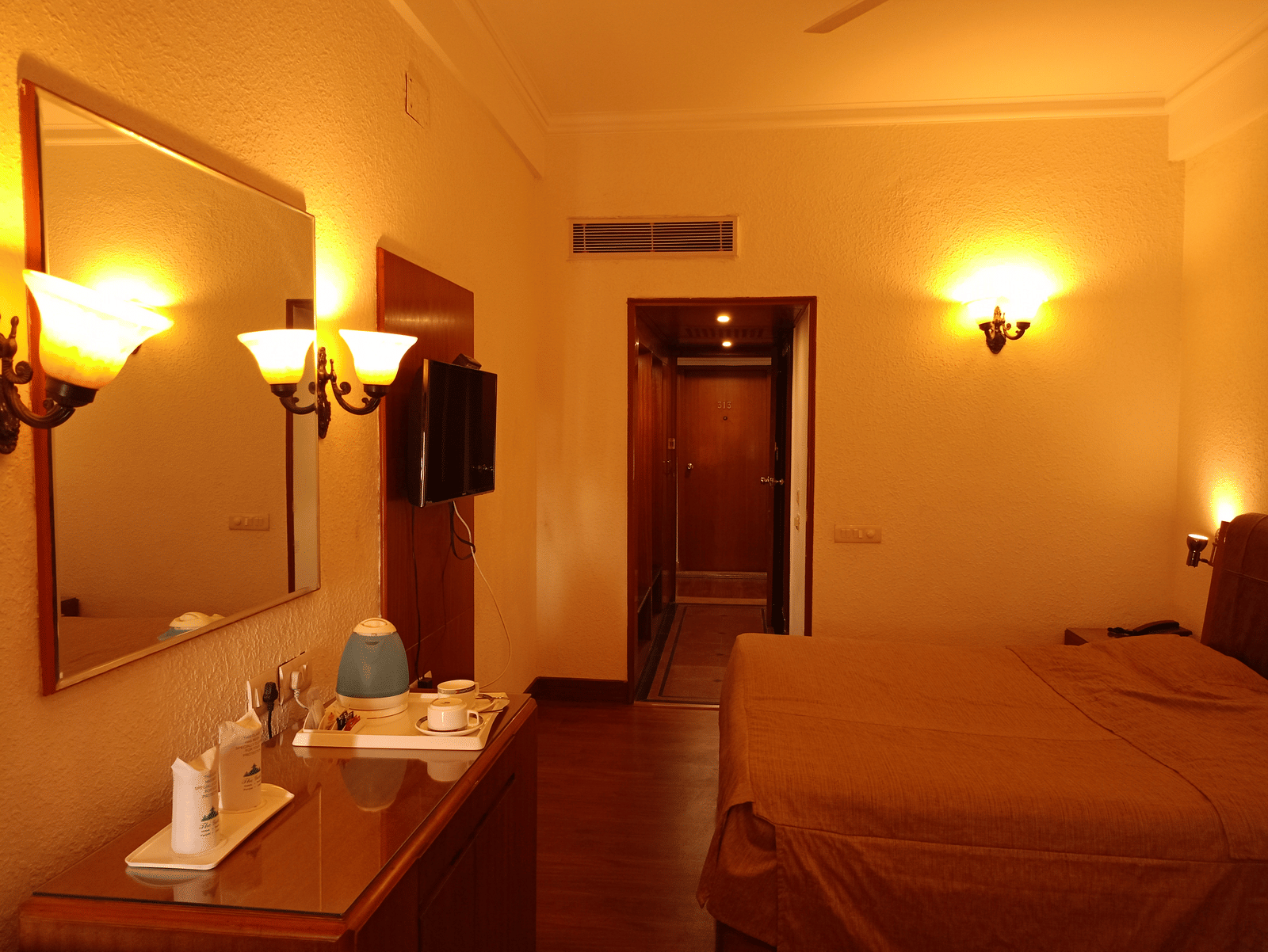 Room interior at Hotel Gold, Panipat with a bed, lamps, desk, and a mirror near the entrance.