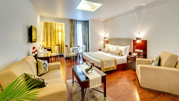 Wide view of a Premium Room at Hotel Hindusthan International Select Bengaluru with a king bed, cream sofas, wooden flooring, coffee table, and skylight.
