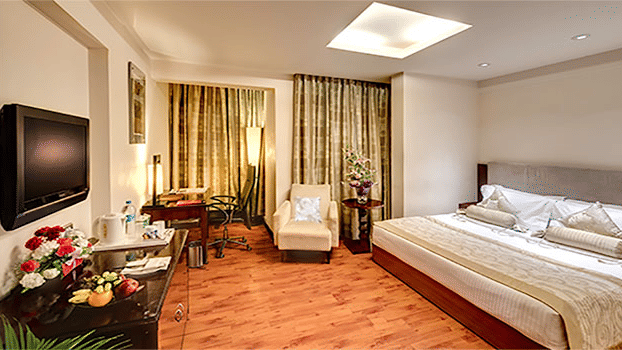 View of a Premium Room at Hotel Hindusthan International Select Bengaluru with a king bed, wall-mounted TV, armchair, skylight, and warm wooden flooring.