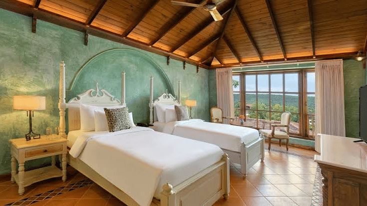 Corner view of a bedroom featuring twin beds kept against a cya-coloured wall, with huge windows overlooking the scenery outside - Golden Tulip Goa Mandrem
