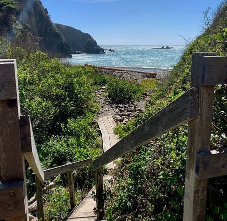 A path descends via steps towards a beach framed by cliffs and vegetation, under a partly cloudy sky.