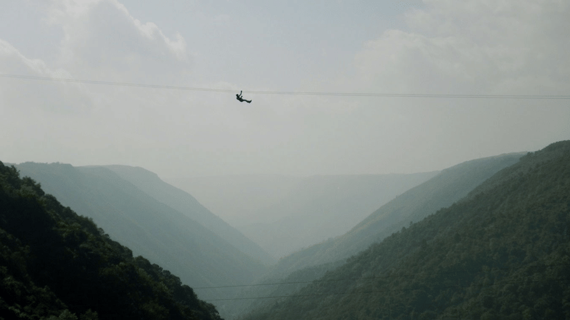 Silhouetted person ziplining across deep forested valley between two green hills in Cherrapunji under hazy pale grey overcast sky.