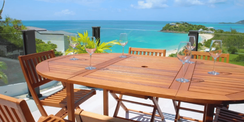 An outdoor dining table set with glasses offers a panoramic view of the sparkling blue Caribbean sea and distant coastline.