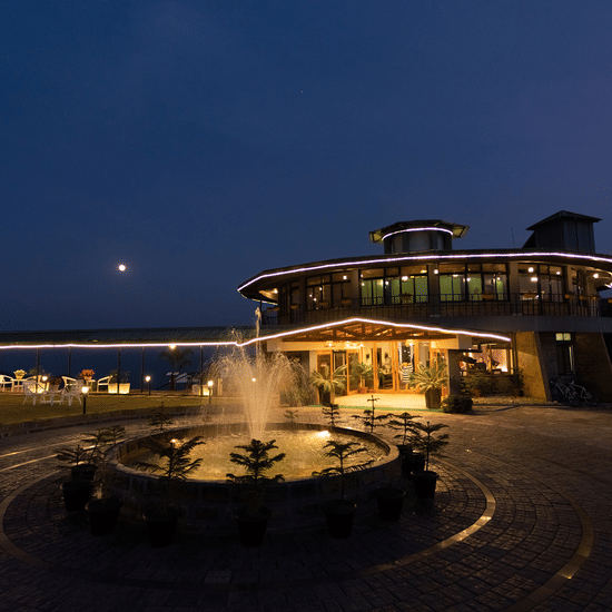 Night facade of Polo Resort Cherrapunji main building outlined in LED lights, with a circular driveway, illuminated fountain and potted conifers in front.