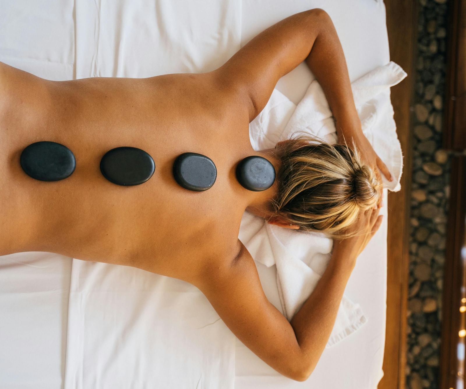 A woman lying down with back with black stones kept over her spine.