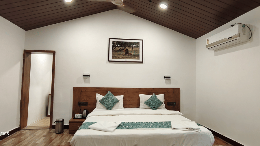 A hotel room with king bed, teal accent pillows, wooden headboard, vaulted ceiling, wall AC, and wildlife artwork above the bed.