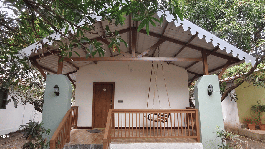 White resort cottage with wooden porch, hanging swing, gabled roof, surrounded by lush mango trees and garden lanterns on pillars.