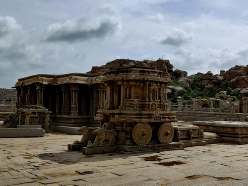 Weathered stone temple ruins in Hampi, one of the popular travelling places in Karnataka, stand across a vast courtyard, reflecting exquisite ancient craftsmanship amid boulder-strewn landscapes and cloudy skies overhead.