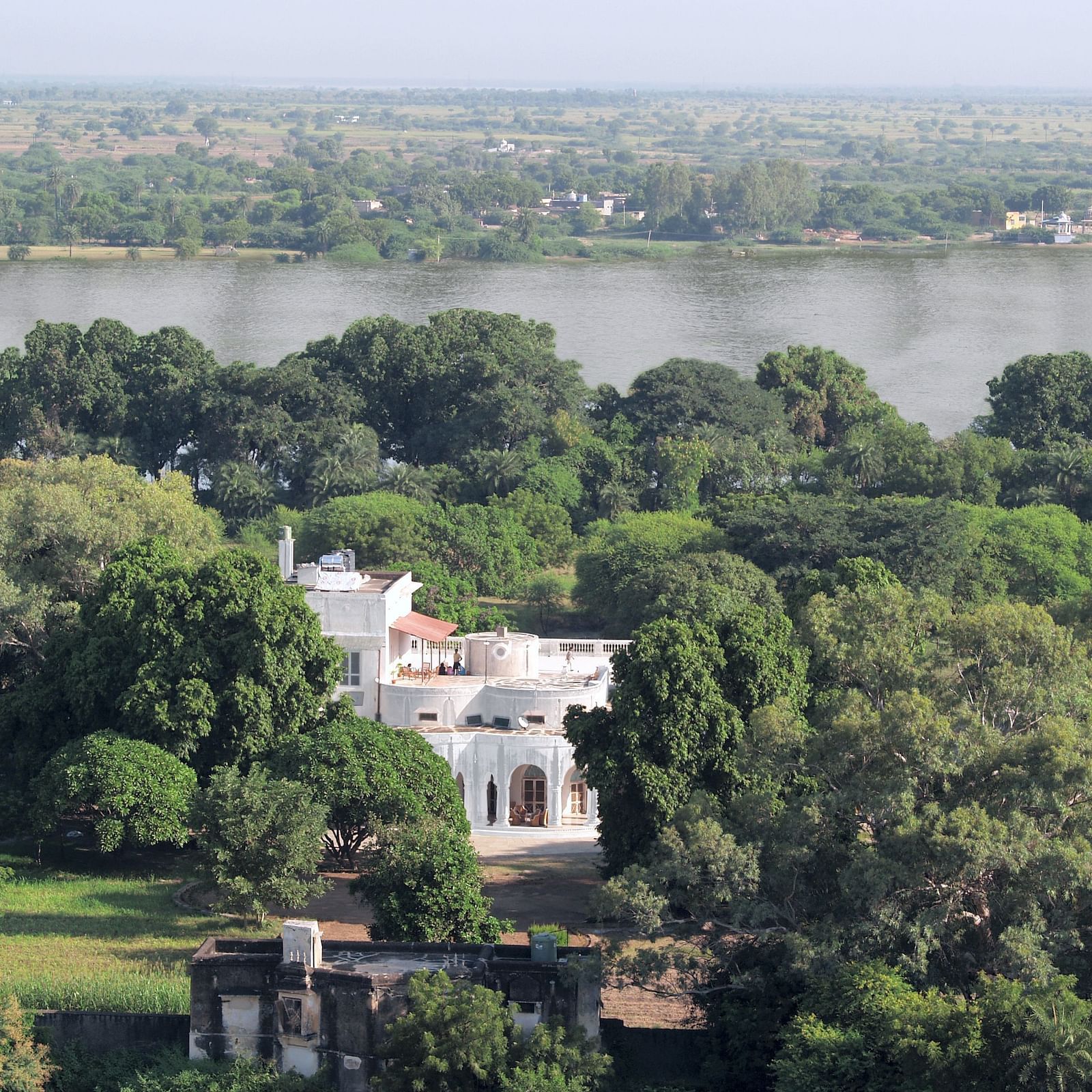 An aerial view of a white building with arched openings, nestled among trees with a wide body of water in the distance- Shahpura Bagh.