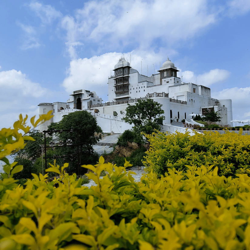 White Sajjangarh Palace on a hilltop with green hedges and cloudy blue skies in the foreground
