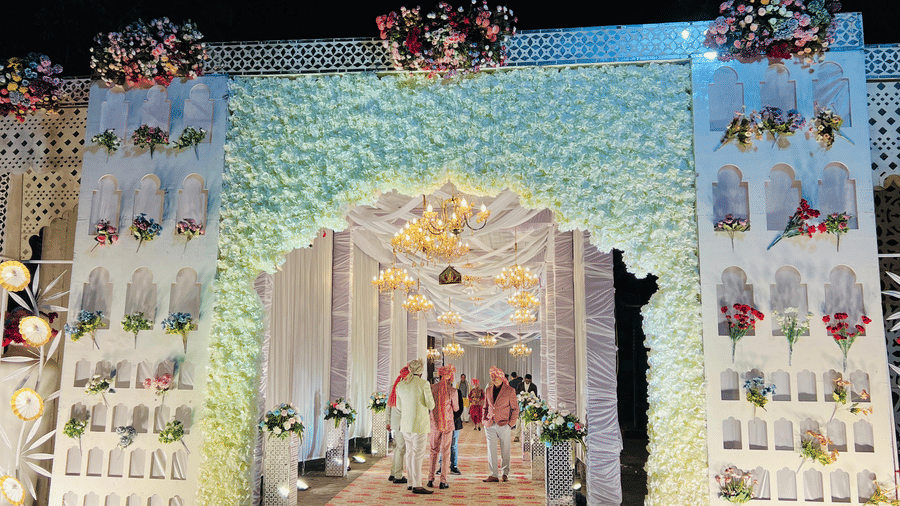 A nighttime view of a wedding aisle covered by a red carpet, leading through a large, white floral archway flanked by decorated pillars | Nandan Resort